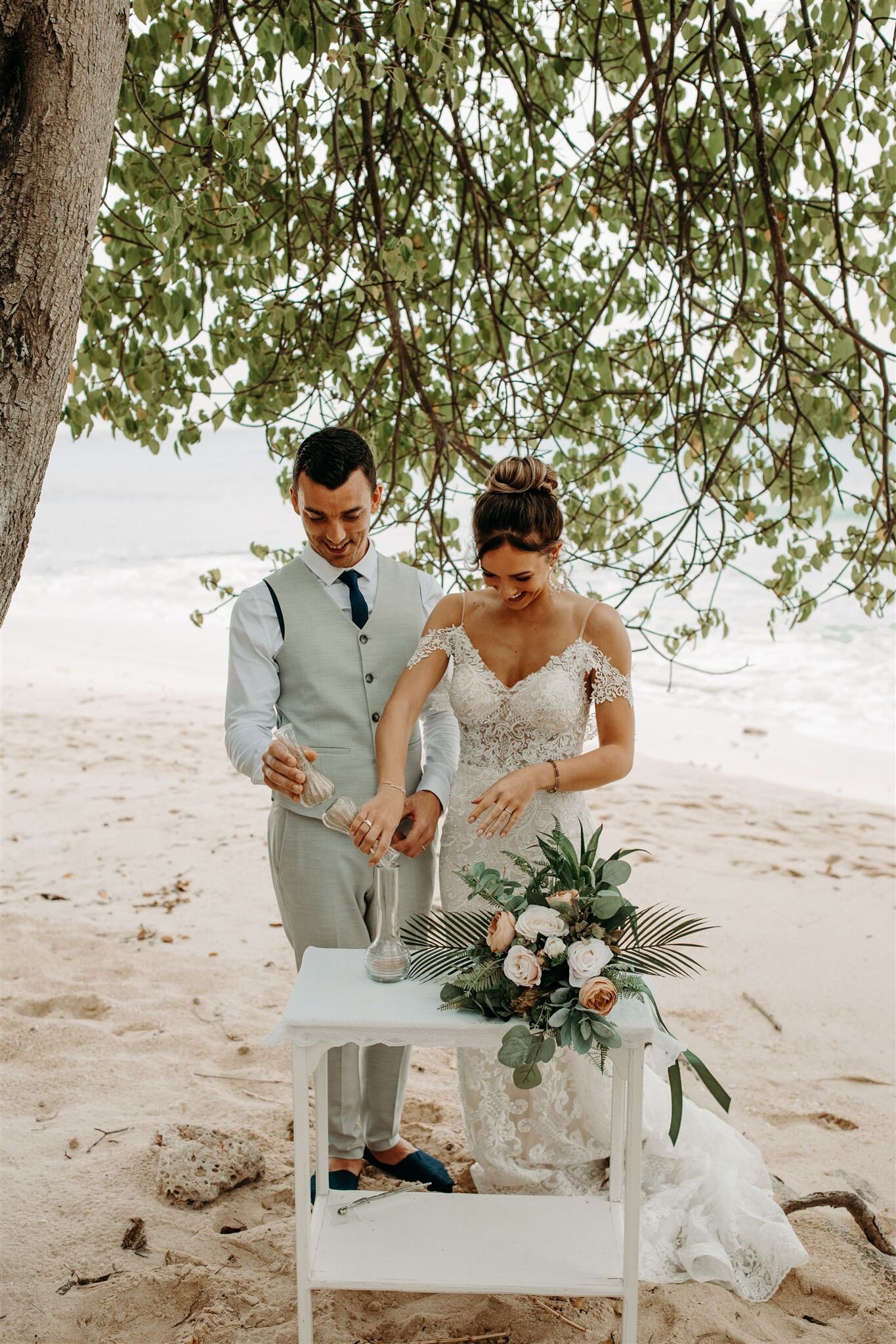 couple joins together pouring sand into a vase as part of their destination elopement beach ceremony. brides bouquet sits on a small white antique table as they smile together.