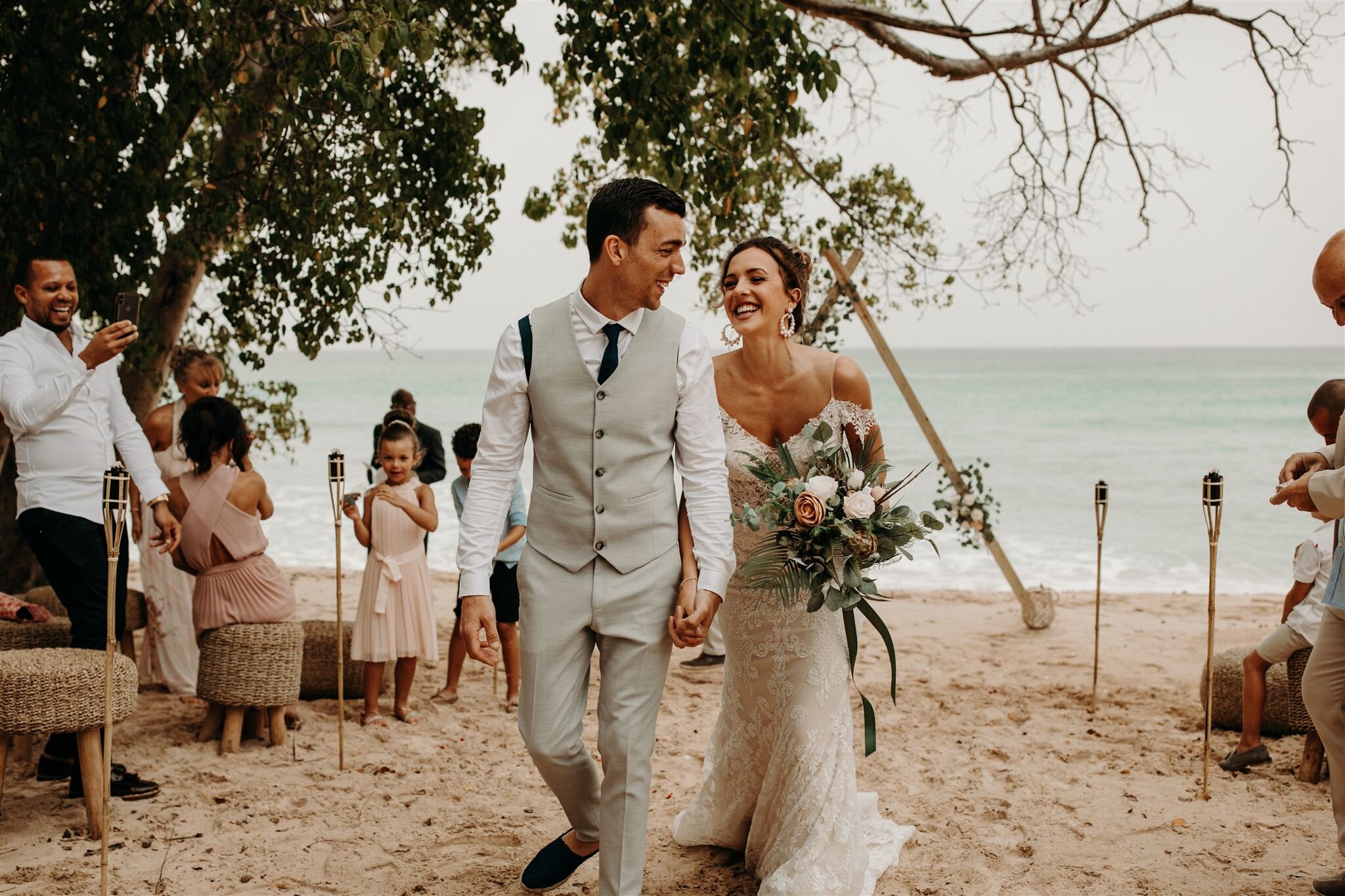 newlyweds laugh and hold hands after boho beach ceremony with friends and family laughing and dancing in the background