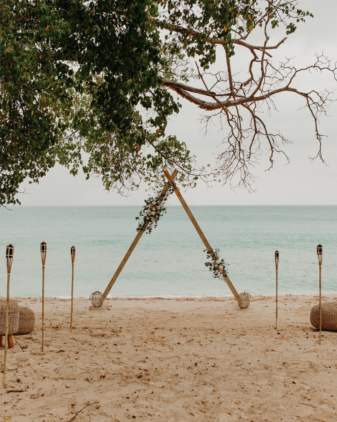 Sustainable wooden triangular arch sits on the beach for a boho wedding ceremony. The arch is accented with florals and tiki torches line the aisle. Behind the tiki torches are boho benches for guests to sit.