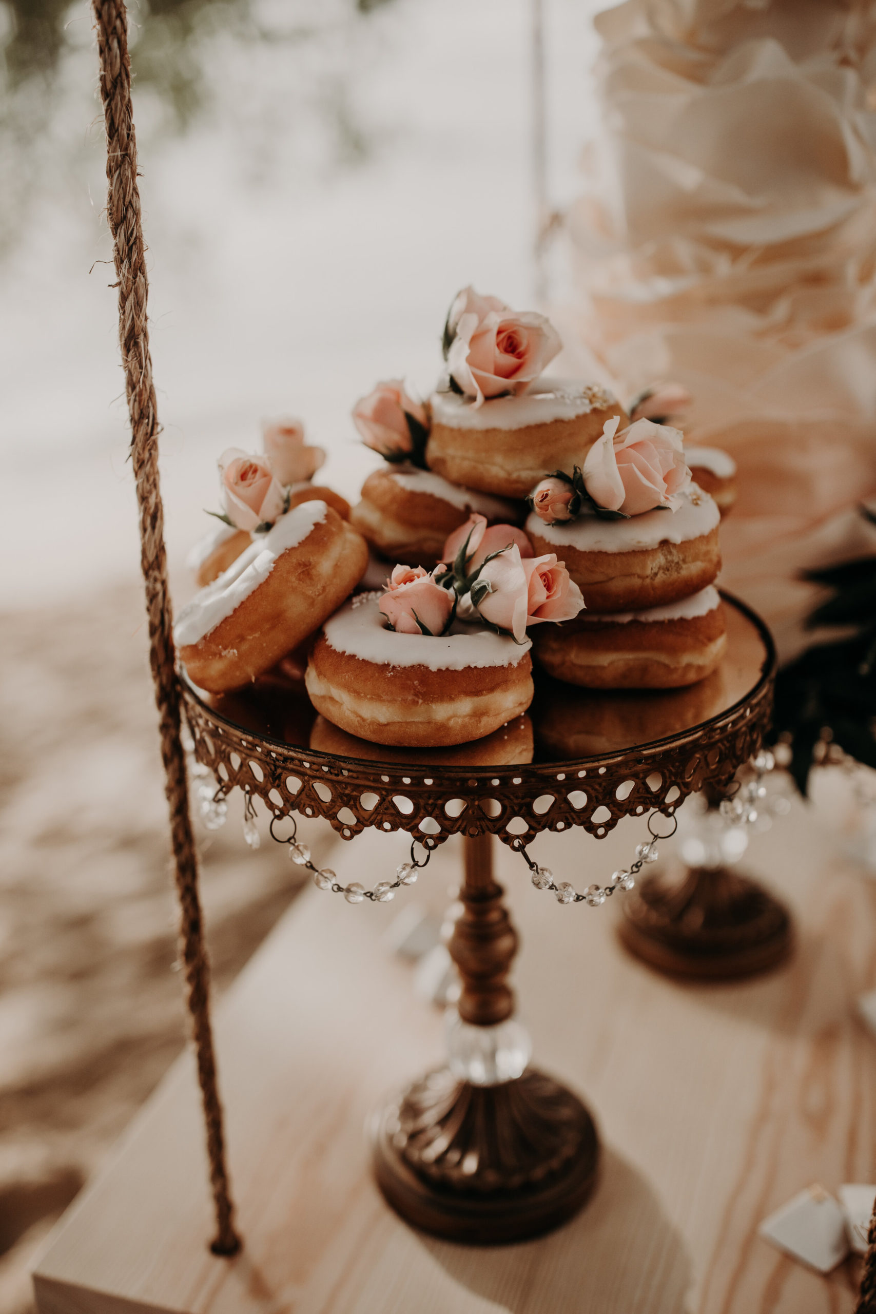 Wedding dessert display piece holding white frosted donuts with pink roses peeking between them as an accent. Out of focus, in the background is a floral tiered wedding cake.